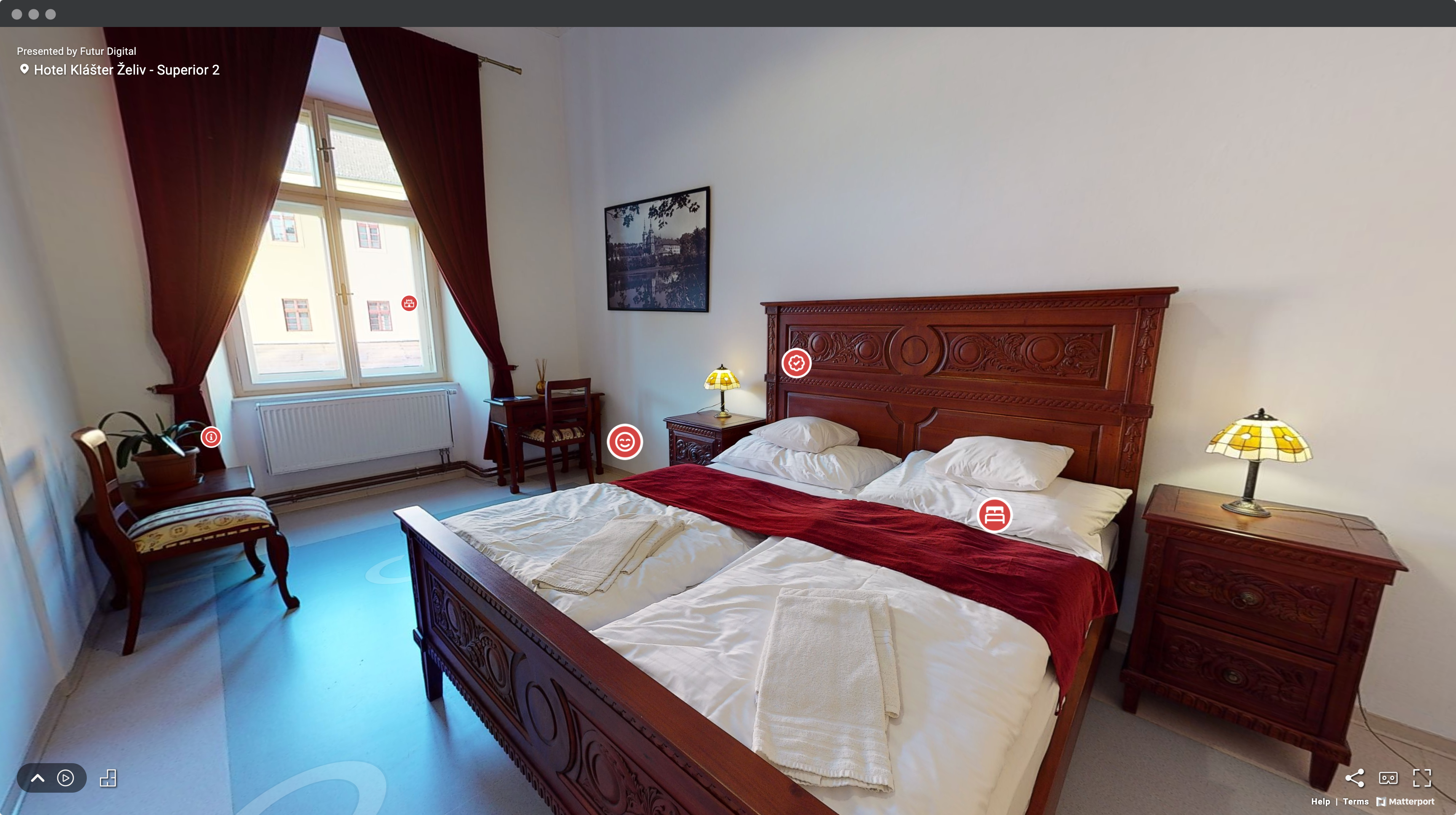 Screenshot okna prehliadača s virtuálnou prehliadkou hotelovej izby v Hoteli Klášter Želiv so záberom na ručne vyrezávanú masívnu drevenú manželskú posteľ s bielymi prikrývkami a červenou Štólou, s nočnými stolíkmi s lampami v Štýle Tiffany, dvomi stolmi a stolićkami a veľkým oknom s červenými závesmi. Označené sú bodyy záujmu.