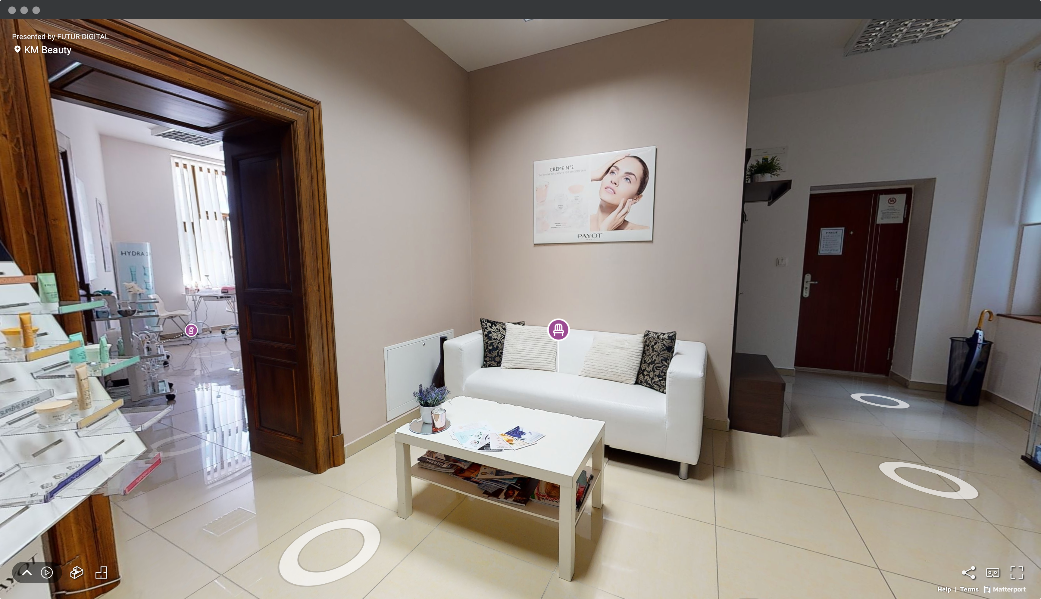 Screenshot okna prehliadača s virtuálnou prehliadkou kozmetického salóna KM Beauty so záberom na vstupnú halu so sedačkou, stolom, vankúšmi a virtínou s kozmetikou.