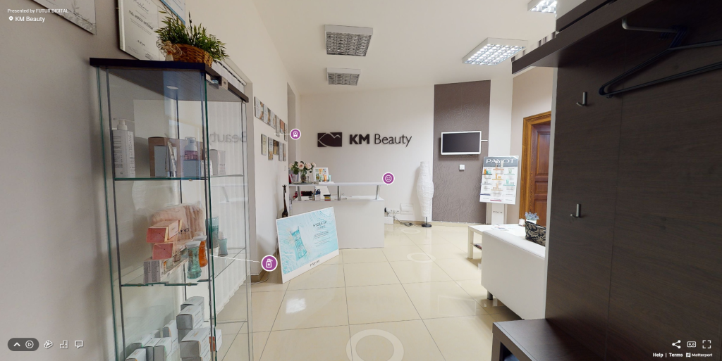 Pohľad na virtuálnu prehliadku beauty salóna KM Beauty so záberom na vstupnú halu s recepciou, sedačkou, stolom, virtínou s kozmetikou a označenými bodmi záujmu.