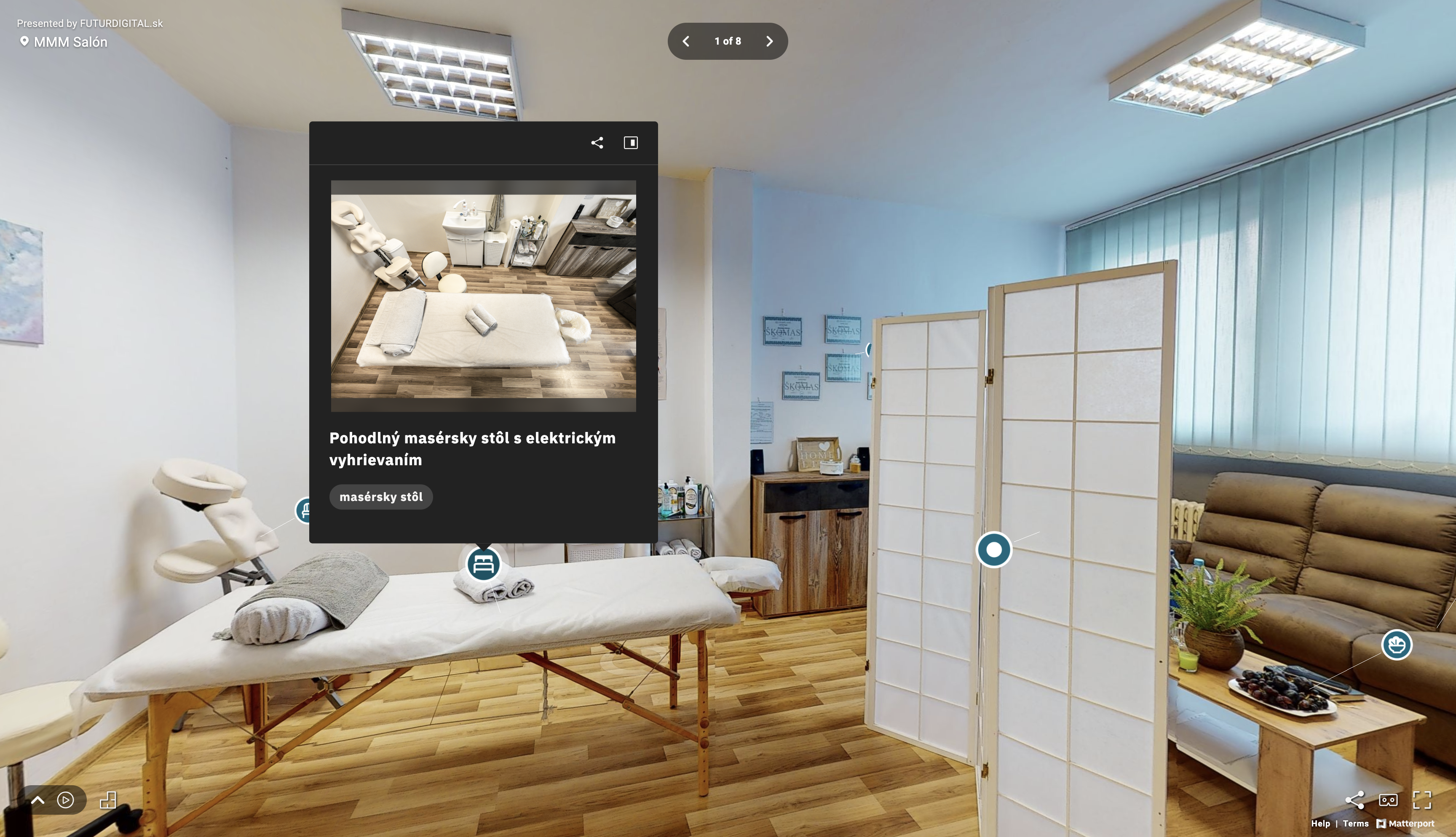 Pohľad na virtuálnu prehliadku masážneho salóna MMM Salón s pohľadom na masážny stôl, paraván, sedaciu súpravu a označený bod záujmu - fotku masáženho stola zhora s popisom vybavenia.