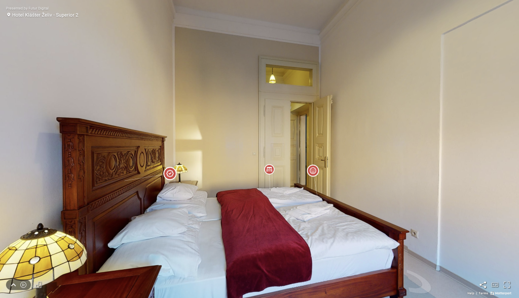 Pohľad na virtuálnu prehliadku hotelovej izby v Hoteli Klášter Želiv s pohľadom na masívnu manželskú posteľ, nočné stolíky s Tiffany lampami a dverami vedúcimi do chodby.
